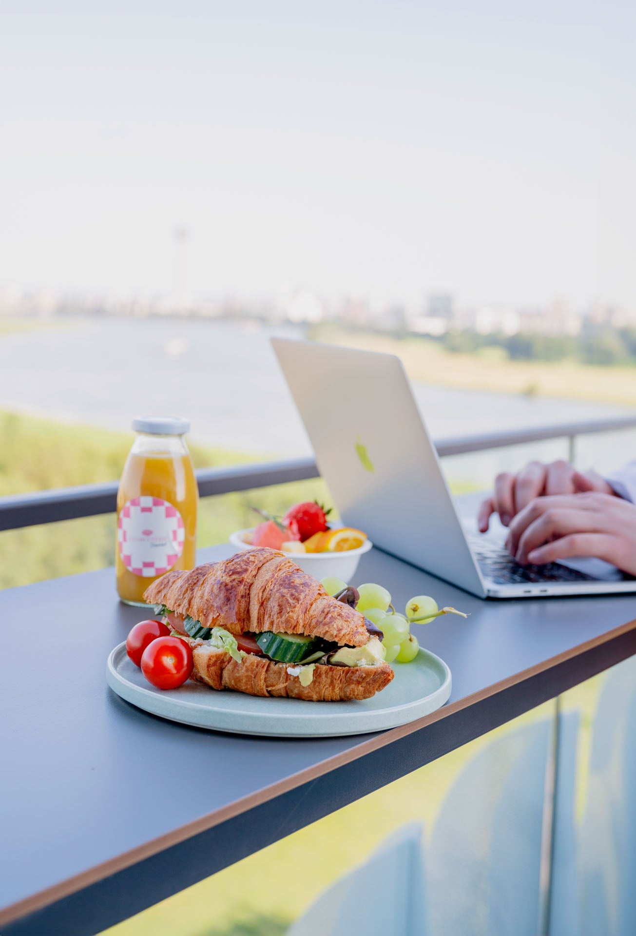 Frisch belegtes Croissant mit Gemüse, Trauben, Tomaten und Orangensaft als Frühstücksbox auf einem Balkon mit Rheinblick – ideal fürs Business-Brunch