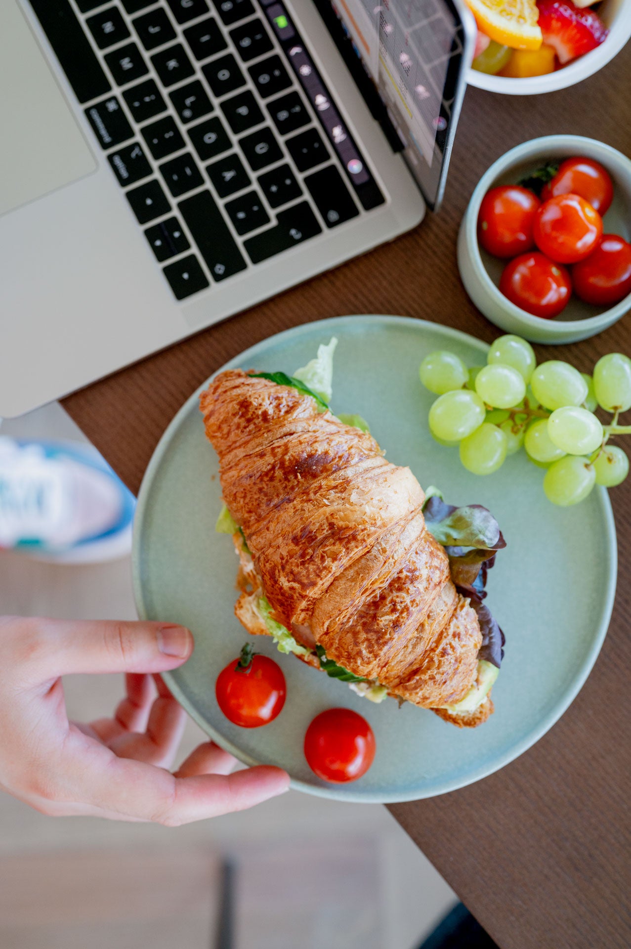 Frühstückscroissant mit Salatfüllung, Kirschtomaten und Trauben neben einem Laptop – perfekte Lunchbox für Meetings oder Homeoffice