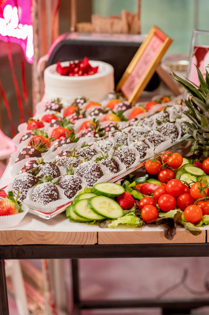Eine köstliche Auswahl an frischen Erdbeeren, getaucht in Schokolade und bestreut mit Kokosraspeln, präsentiert neben einer Platte mit frischem Gemüse wie Tomaten und Gurken, alles elegant angeordnet auf einem Catering-Tisch bei einer Veranstaltung.