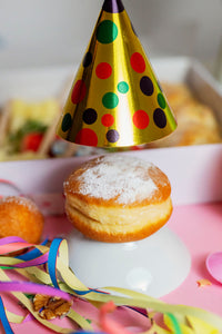 Berliner mit Puderzucker, dekoriert mit einem bunten Partyhütchen, umgeben von Luftschlangen und Konfetti. Ein fröhliches Arrangement auf einem pinken Hintergrund, perfekt für Karnevalsfeiern in Düsseldorf.