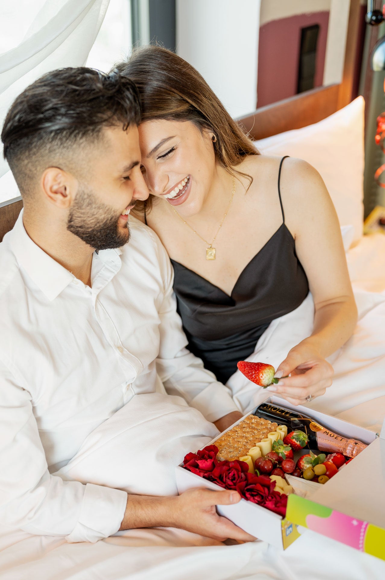 Paar in romantischer Stimmung, das im Bett eine Valentinstagsbox mit frischen Erdbeeren, Trauben, Käse und Moët Champagner genießt. Die perfekte Szene für einen liebevollen und unvergesslichen Valentinstag.