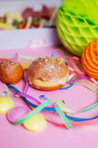 Karnevalsbox Düsseldorf mit einem glasierten Berliner, einer süßen Kugel, buntem Konfetti, Luftschlangen und knackigen Walnüssen auf einem pinken Hintergrund. Im Hintergrund sind Käsewürfel, frisches Gemüse, Brot und Süßigkeiten aus der Box zu sehen – perfekt für Karnevalsfeiern.