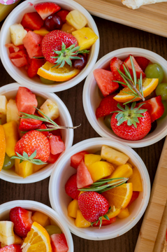 Mini fruit bowls including strawberries, grapes, oranges, watermelon, pineapple, mango and peach. Topped with rosemary, placed on a wooden board,