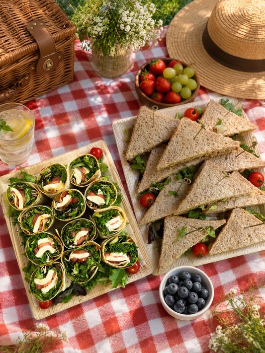 Picnic mat red white with some fruits and some finger food like sandwiches and wraps. 