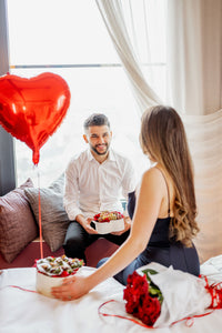 Paar in einem romantisch dekorierten Zimmer mit roten Luftballons; der Mann überreicht der Frau eine Valentinstagsbox mit Macarons.