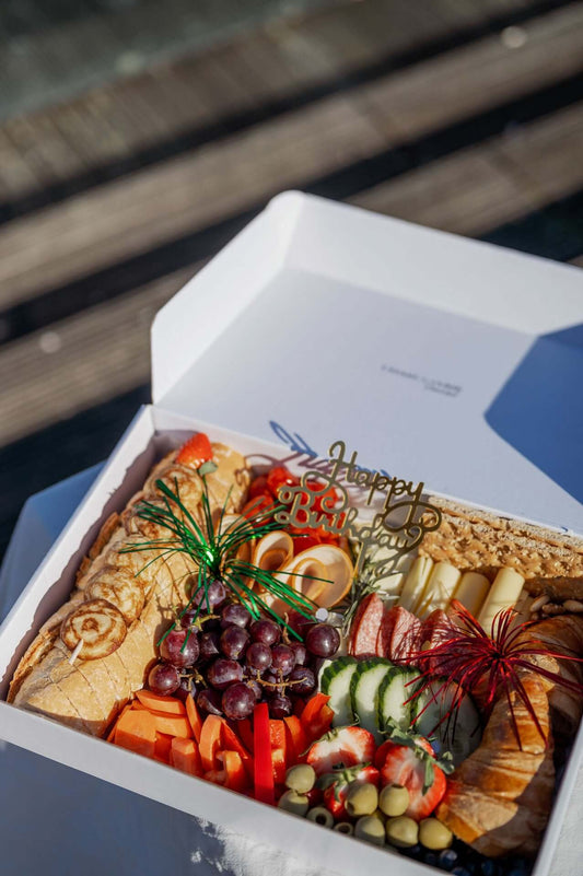 Eine offene Geschenkbox mit einer köstlichen Auswahl an Snacks, perfekt für eine Geburtstagsfeier. Die Box beinhaltet frisch geschnittenes Obst, wie Trauben, Erdbeeren und Melone, zusammen mit Gemüsesticks, Käse, Wurstscheiben und einem knusprigen Baguette. In der Mitte der Snacks steckt eine goldene Dekoration mit der Aufschrift "Happy Birthday", die festlich in die Szene integriert ist.