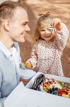 Lade das Bild in den Galerie-Viewer, Ein fröhlicher Vater und seine kleine Tochter genießen ein Picknick, wobei das Mädchen stolz eine Traube hochhält. Die beiden befinden sich im Freien, und vor ihnen ist eine Picknickbox mit einer vielfältigen Auswahl an Snacks zu sehen, darunter Käse, Wurstwaren und frisches Obst. Dieses Bild strahlt familiäre Verbundenheit und die Freude an gemeinsamen Aktivitäten in der Natur aus.
