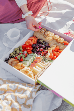 Frühstücksbox mit frischen Zutaten wie Obst, Gemüse, Croissants, Käse und Brot bei einem sonnigen Picknick. Eine Frau im weißen Pullover und rosa Rock greift nach einem Snack aus der liebevoll arrangierten Box. Ideal für Frühstück im Freien oder gemütliche Picknick-Momente.