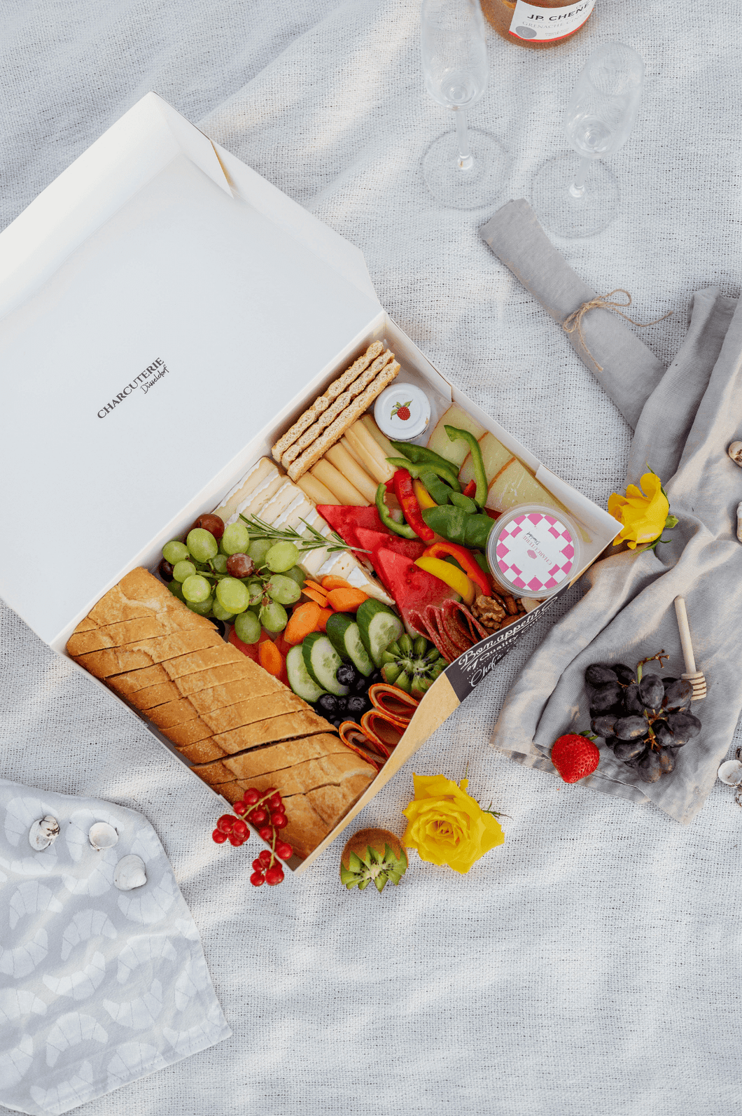 A charcuterie Box is laying on the floor. The food box has different ingredients inside it such as Bread, cheese, meat, fruits, vegetables, dips, nuts and more. The box is beautifully decorated with some decoration beside it.