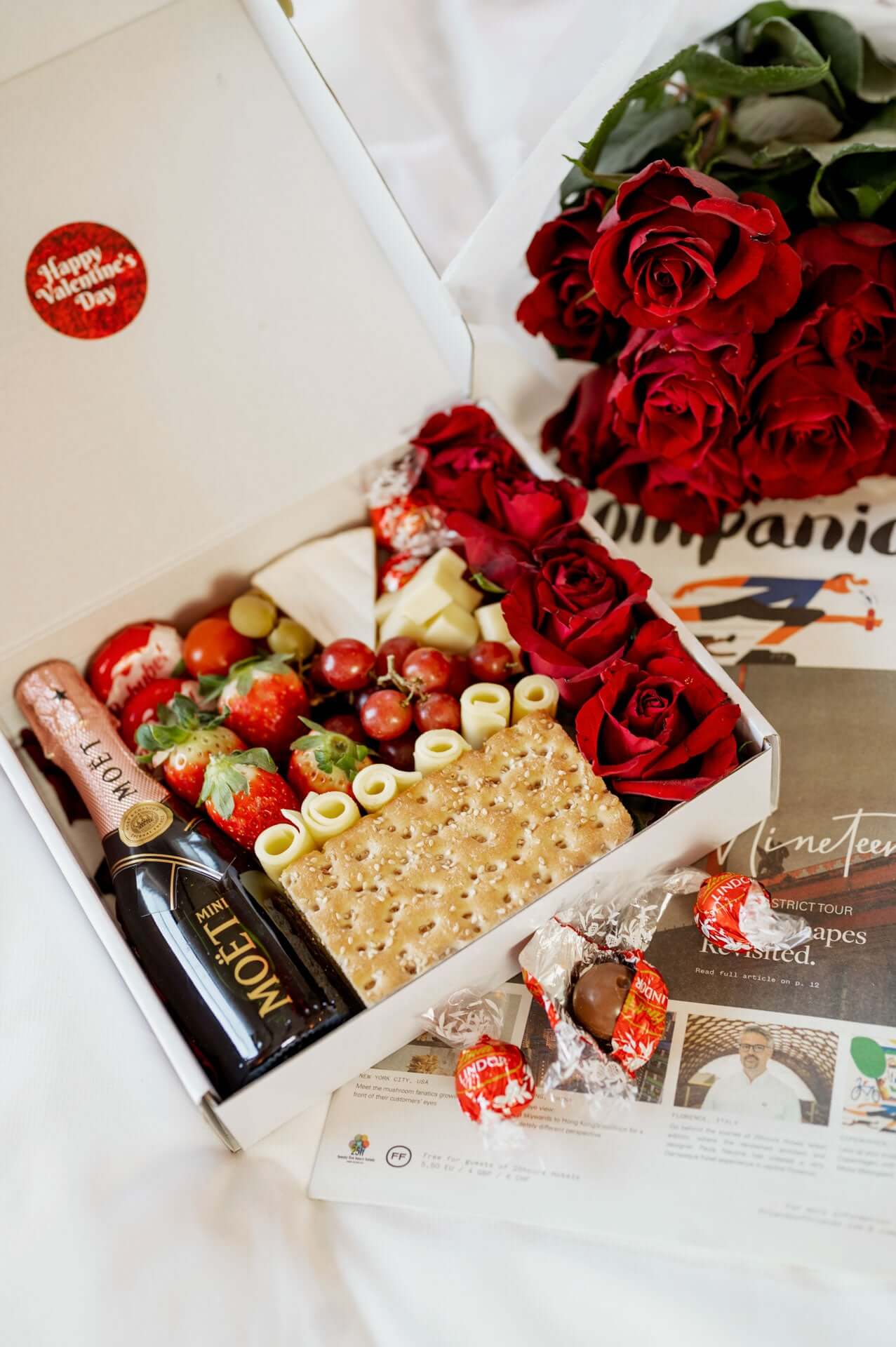 Valentinstagsbox mit einer Auswahl an roten Rosen, frischen Erdbeeren, Trauben, Käse, Crackern und einer Mini-Flasche Moët Champagner. Stilvoll präsentiert mit Pralinen und liebevollen Details, perfekt für einen romantischen Anlass.
