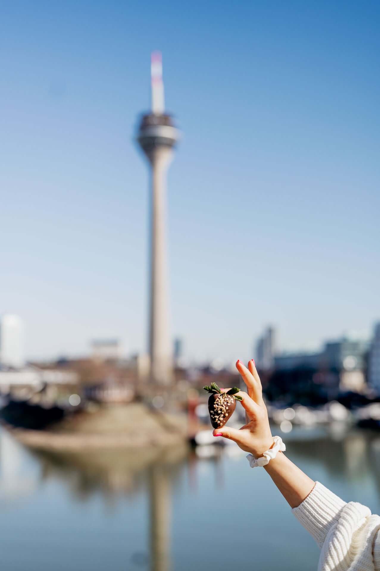 Hand hält eine mit dunkler Schokolade überzogene Erdbeere vor der Skyline von Düsseldorf, mit dem markanten Rheinturm im Hintergrund. Perfekte Kombination aus Genuss und urbanem Flair an einem sonnigen Tag.