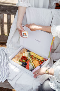 A picture of an elegantly dressed person holding a 'I ♥ MUM' cookie over a beautifully arranged box filled with a luxurious selection of picnic foods, including various cheeses, fruits, and bread. The image conveys a sense of preparation for a sophisticated Mother's Day celebration or a special occasion picnic, emphasizing thoughtfulness and care in the presentation.