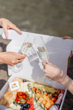 Load image into Gallery viewer, Two hands clinking champagne flutes above a box filled with an elegant assortment of gourmet foods, including cheese, fruits, and charcuterie, near a body of water. The setting suggests a celebratory outdoor event, possibly a picnic or a social gathering, with a focus on shared enjoyment and fine dining in a casual yet sophisticated environment.
