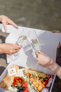Two hands clinking champagne flutes above a box filled with an elegant assortment of gourmet foods, including cheese, fruits, and charcuterie, near a body of water. The setting suggests a celebratory outdoor event, possibly a picnic or a social gathering, with a focus on shared enjoyment and fine dining in a casual yet sophisticated environment.