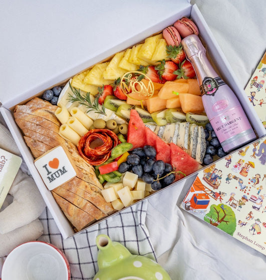 An appealing picnic spread showcasing a variety of fruits like strawberries, blueberries, pineapple, and watermelon, alongside assorted cheeses, bread, and charcuterie. Accents like a 'Best Mom' strawberry, 'I ♥ MOM' cookie, and a small bottle of Brut Dargent wine add a personal touch, suggesting a Mother's Day theme. The picnic is complemented by a children's book and a whimsically illustrated cover, hinting at a family-friendly outing.