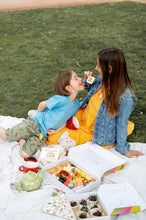 Cargar imagen en el visor de la galería, A candid moment captured of a mother and child enjoying a picnic, with the child playfully biting into a cookie held by the mother. They are seated on a blanket adorned with a teapot, cups, and boxes of gourmet treats, indicative of a leisurely outdoor family activity. Their relaxed postures and smiles suggest a joyful and comfortable bond, set on a grassy field which adds a natural and fresh atmosphere to the scene.
