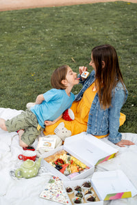A candid moment captured of a mother and child enjoying a picnic, with the child playfully biting into a cookie held by the mother. They are seated on a blanket adorned with a teapot, cups, and boxes of gourmet treats, indicative of a leisurely outdoor family activity. Their relaxed postures and smiles suggest a joyful and comfortable bond, set on a grassy field which adds a natural and fresh atmosphere to the scene.
