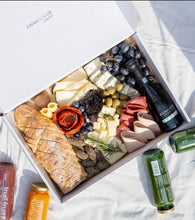 Cargar imagen en el visor de la galería, An overhead view of a gourmet picnic box with compartments filled with an assortment of snacks: dark chocolate-covered strawberries, a variety of cheeses, black grapes, bread, salami, and crackers, alongside a bottle of SCAVI &amp; RAY alcohol-free sparkling wine. Adjacent to the box are bottles of True Fruits smoothie, adding a healthy, refreshing option to the indulgent spread.
