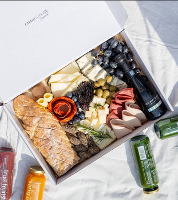 An overhead view of a gourmet picnic box with compartments filled with an assortment of snacks: dark chocolate-covered strawberries, a variety of cheeses, black grapes, bread, salami, and crackers, alongside a bottle of SCAVI & RAY alcohol-free sparkling wine. Adjacent to the box are bottles of True Fruits smoothie, adding a healthy, refreshing option to the indulgent spread.